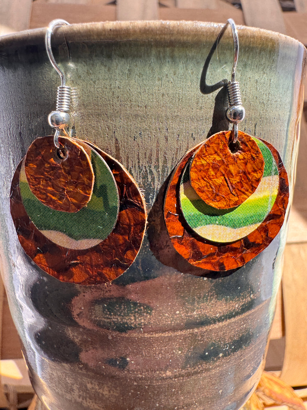 Handmade layered cork and fabric earrings in warm brown, green, and gold tones displayed on a ceramic vase