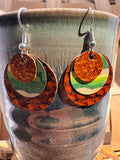 Handmade layered cork and fabric earrings in warm brown, green, and gold tones displayed on a ceramic vase